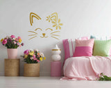 Wall decal of a simple cat face with closed eyes, whiskers, and a floral design on one ear. The design is cut from gold vinyl with no background and is displayed above a bed with pink and green pillows and baskets of flowers.