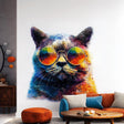 Printed wall decal of a colorful cat wearing large reflective sunglasses. The decal is cut along the shape of the cat and applied to a white wall, shown above a sofa with orange and blue cushions.