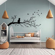 Vinyl wall decal of a cat sitting on a tree branch with flying birds, cut in solid black and applied to a pale green wall above a child&#39;s bed in a cozy, modern nursery.
