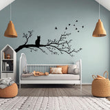 Vinyl wall decal of a cat sitting on a tree branch with flying birds, cut in solid black and applied to a pale green wall above a child&#39;s bed in a cozy, modern nursery.