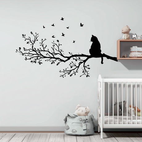 Vinyl wall decal of a cat sitting on a detailed tree branch with flying birds. The decal is solid black with no background and is applied above a crib in a soft-toned nursery.