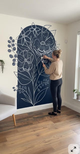 Woman applying a large floral wall decal to a white wall. The design features hand-drawn white flowers and leaves on a navy blue arched background. The decal is printed on vinyl and cut to shape, blending naturally into the room’s decor.