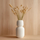 Tall 3D printed white geometric vase with ribbed texture, holding dried grass stems, displayed against a light wood background