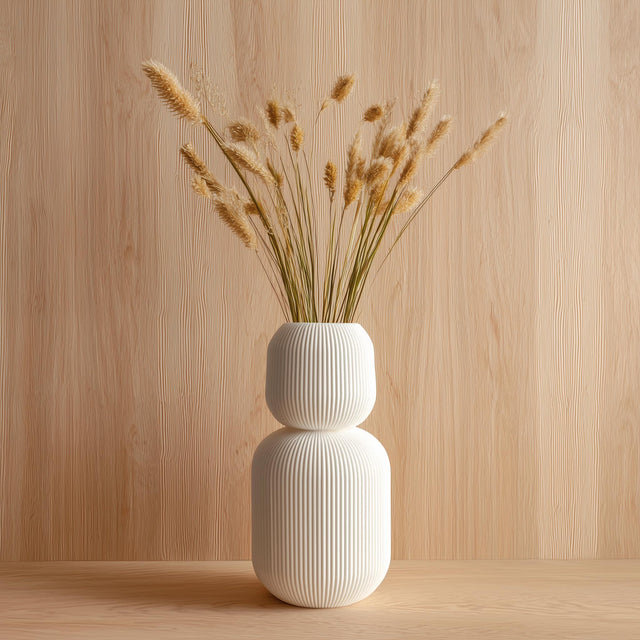 Tall 3D printed white geometric vase with ribbed texture, holding dried grass stems, displayed against a light wood background