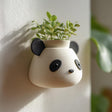 3D printed wall-mounted planter shaped like a cute panda head in matte speckled PLA, holding a small green plant against a light wall in soft natural light.