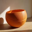 Orange 3D printed bowl shaped like a sliced basketball made from matte PLA, standing on a wooden surface in soft natural light.