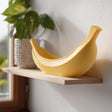 Yellow 3D printed banana-shaped bowl made of matte PLA plastic standing on a light wooden shelf near a window in soft natural daylight.