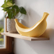 Yellow 3D printed banana-shaped bowl made of matte PLA plastic standing on a light wooden shelf near a window in soft natural daylight.