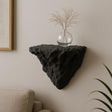 A black 3D printed wall-mounted shelf with a rugged, rock-like texture and triangular shape, displayed in a minimalist interior. The shelf holds a round glass vase with dried ornamental grass, and is mounted above a beige couch against a soft-toned wall.