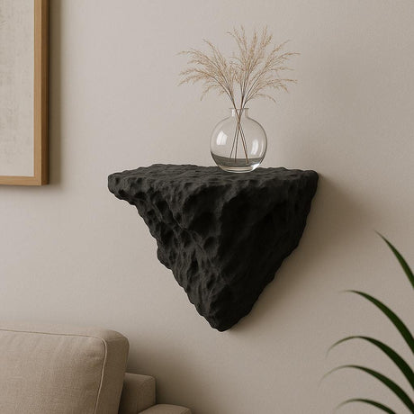 A black 3D printed wall-mounted shelf with a rugged, rock-like texture and triangular shape, displayed in a minimalist interior. The shelf holds a round glass vase with dried ornamental grass, and is mounted above a beige couch against a soft-toned wall.