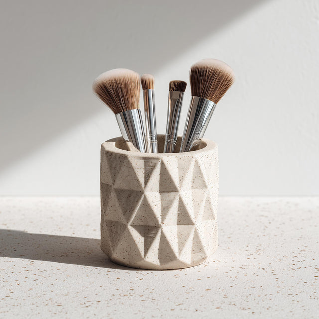 A beige 3D printed geometric cup holding makeup brushes, shown on a speckled surface with natural light. The cup features a faceted, textured design and matte finish.