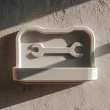 3D printed wall-mounted tool shelf with a shallow tray and embossed wrench icon on the back panel, matte light PLA finish, fixed on a textured workshop wall.