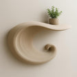 A beige 3D printed floating shelf in the shape of a curling wave, mounted on a light wall and holding a small potted plant on top.