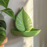 3D printed leaf-shaped wall shelf made of matte green PLA plastic, mounted on a wall near plants, modern botanical home decor accessory