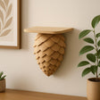 A wall-mounted shelf designed to resemble a pinecone, 3D printed in matte tan material. The shelf has overlapping scale-like layers forming a conical shape pointing downward, with a flat surface on top displaying minimalist decor. It is mounted on a neutral-colored wall above a wooden surface, surrounded by potted plants and natural-toned home accents.