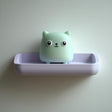 3D printed wall shelf with a small cube cat character sitting in a rounded tray-like ledge, made of matte PLA plastic on a light-colored wall.