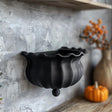 3D printed wall shelf shaped like a black witch cauldron with wavy rim, matte PLA texture, mounted on a textured grey wall in a cozy autumn interior.