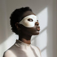 Adult person wearing a minimalist white 3D printed eye mask, standing in soft natural light against a neutral interior background.