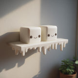 3D printed wall shelf with a dripping edge and two marshmallow-like cube characters with faces, made of matte PLA plastic and mounted on an interior wall in natural light.