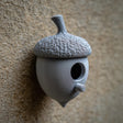 3D printed acorn-shaped birdhouse with round entrance and small cylindrical perch made of matte PLA plastic, mounted on a textured exterior wall.