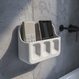 3D printed white wall-mounted organizer shelf with two upper compartments for phone and remote and three small front pockets, installed above a bathroom sink on a textured grey wall.