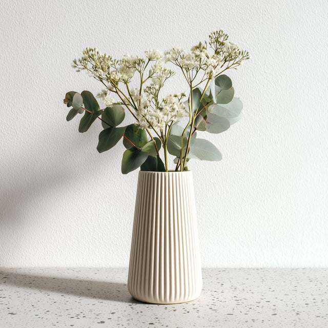 A tall, cream-colored 3D printed origami vase with vertical ribbed lines, holding white dried flowers and eucalyptus stems, placed on a speckled countertop.
