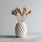 A white geometric 3D printed vase with a diamond-textured surface, holding dried beige flowers, shown on a gray concrete surface against a light background.