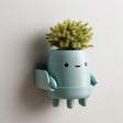 3D printed matte PLA wall planter shaped like a cute character with little legs, a round body used as a pot and a small side tray, mounted on a light wall.
