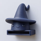 3D printed matte PLA wall shelf shaped like a cute wizard hat character with a small open storage cubby, mounted on a light wall.