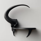 3D printed matte PLA wall shelf with a stylized ram skull and long curved horn supporting a small floating ledge on a light-colored wall.