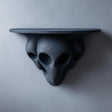 3D printed dark gray wall shelf with three elongated alien heads forming a sculptural support and a flat floating ledge on a light textured wall.