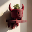 Wall-mounted 3D printed planter shelf featuring a cute red devil-shaped pot holding a small succulent, designed as playful fantasy wall decor.