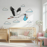 A flat-style illustration on a white background showing a large white stork with black-edged wings in flight. The stork carries a small, smiling baby in a light blue cloth bundle hanging from its orange beak, surrounded by simple white clouds.