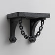 3D printed matte black wall shelf with two curved brackets connected by a decorative hanging chain, mounted on a light textured wall.