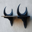 3D printed dark fantasy wall shelf supported by two curved dragon horn sculptures, mounted on a textured light wall with a slim floating ledge.