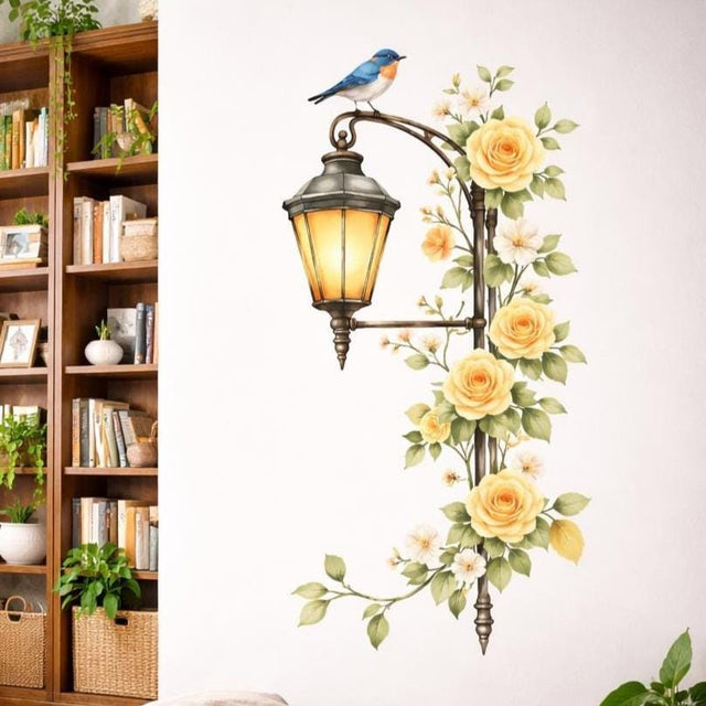 A vertical watercolor illustration of an antique dark metal street lamp on a white background. The lamp is lit with a warm yellow glow and is entwined with a vine of pale yellow roses and small white flowers. A bluebird with an orange breast sits on the curved arm of the lamp.