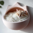 3D printed crown-shaped wall mounted cat bed in matte blush PLA with a fluffy cream cat sleeping inside, installed on a textured light wall.
