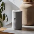 3D printed minimalist cylindrical pet ashes urn with a raised paw print emblem and fitted lid, displayed on a tabletop in soft natural light.