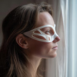 3D printed minimalist contour eye mask with flowing side cutout panels, worn in side profile near a window with soft daylight, visible print texture.