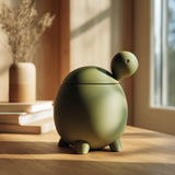 3D printed turtle-shaped pet ashes urn in matte olive green PLA with visible print texture, sitting on a wooden table near a bright window with warm decor.