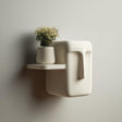 3D printed minimalist face-inspired wall shelf with a compact floating ledge and sculptural side detail, holding a small vase, mounted on a clean wall in soft light.