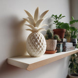 3D printed geometric pineapple memorial urn in matte PLA displayed on a floating wall shelf beside indoor plants in a modern interior.