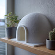 3D printed white igloo dome decor with arched entrance displayed on a wooden shelf beside small plants in a modern interior.