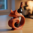 3D printed fox-shaped wine bottle holder in matte orange PLA holding a wine bottle on a wooden kitchen table with warm lighting.
