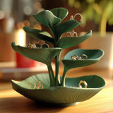 Green botanical jewelry holder with layered leaf-shaped trays holding rings and earrings on a wooden surface in warm natural light.