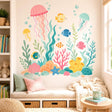 Pastel underwater illustration with three jellyfish, five colorful fish, floating bubbles, seaweed, coral, a blue starfish, pink shell, yellow sea sponges, and rounded ocean rocks on a white background.
