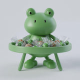 A close-up photograph of a 3D printed cartoon frog figurine standing behind a small round serving table tray filled with wrapped candies. The cute frog features a smooth rounded head, expressive black eyes, and stubby legs supporting the round snack tray.