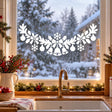 White Christmas window garland with three bells beneath a bow and snowflake, framed by foliage, ornaments and smaller bows.